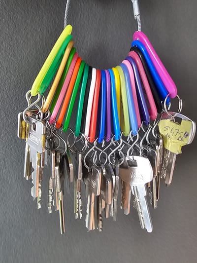 A bunch of keys, with colored plastic tags attached, hangs from a wire against a dark gray textured wall. The keys are made of silver-colored metal and vary in size and shape. Some keys have embossed markings such as "ABUS" and "IKX". The colored tags are arranged in a rainbow-like spectrum of yellow, green, pink, orange, red, purple, blue, black, and magenta. One key has a yellow tag with handwritten numbers and letters that appear to be "547-18" and "15". The lighting suggests an indoor environment, possibly a public building or a storage area. The image is a close-up shot focused on the keys and their tags. There are no people visible in the image. The location is described as being in Darmstadt, Germany.
