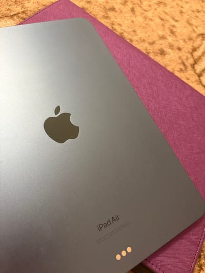 The media file shows a close-up, top-down view of the back of a gray iPad Air tablet. The iPad is resting on a textured, light brown surface, possibly a blanket or rug, and partially overlaps a reddish-purple folio or case. The Apple logo is prominently displayed in the center of the iPad's back. In the upper right corner of the iPad's back, there is small, engraved text that reads "iPad Air" and additional details about its design and manufacturing, including "Designed by Apple in California Assembled in China" and model and serial numbers. Three small, circular dots are also visible near the text. The lighting suggests an indoor setting, and there are no people or active events depicted.