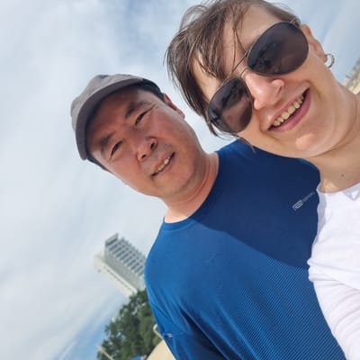 In Gangneung-si, South Korea, a selfie shows two individuals outdoors. A man wearing a gray baseball cap and a blue shirt faces the camera, his head tilted upwards. To his right, a woman wearing sunglasses and a white top also looks at the camera, smiling. The background features a bright sky with clouds, and a tall building is visible in the distance, along with some trees. The scene appears to be at ground level, likely a public outdoor space.