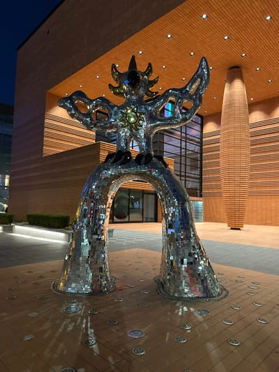 A reflective mosaic sculpture stands in an urban setting. The sculpture is a creature-like form with a sun-like design on its chest and a spiked head. It appears to be standing on two legs. The ground is tiled, with small circular lights embedded in the surface. A brick building with a curved feature in the upper part is behind the sculpture. The building also has large windows. The overall lighting suggests it is dusk or night.