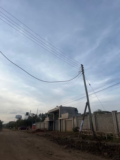 The image displays an outdoor scene in Gitothua ward, Kenya, under a cloudy sky. An unpaved dirt road is visible in the foreground and extends into the distance, bordered by dark soil piles. A prominent wooden utility pole with a diagonal wooden brace stands on the right side of the road, supporting numerous electrical power lines that crisscross the upper portion of the frame. To the right of the utility pole, a large, unpainted concrete structure, consistent with a building under construction, is situated behind a perimeter fence constructed from light grey precast concrete panels. Wooden planks are propped against this fence near the pole. Further along the dirt road to the left, a dark red vehicle, resembling a hatchback, is parked. In the background on the left, two elevated water tanks on metal support structures are visible above a tree line. No individuals are present in the scene.