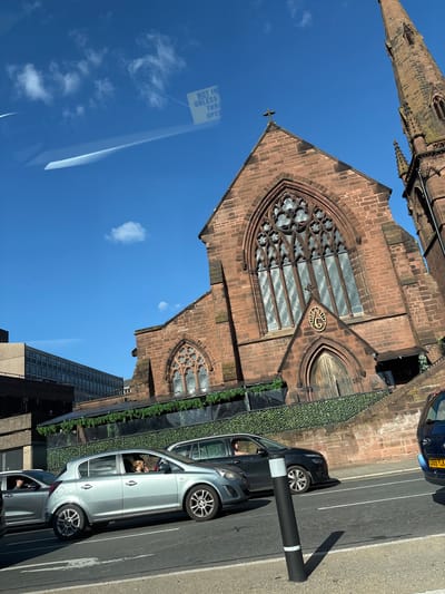 The image captures a view of the ruins of the old Coventry Cathedral in Chester, United Kingdom, taken from inside a vehicle. The cathedral's partial spire and the main section of its facade with a large, ornate window are visible against a clear blue sky with some cloud cover.
In the foreground, there are several cars parked alongside the road. One silver car appears to be occupied, and a black bollard stands beside the road. The scene is in an urban environment, with parts of modern buildings visible in the background. The image suggests a blend of historical ruins juxtaposed with contemporary urban life.