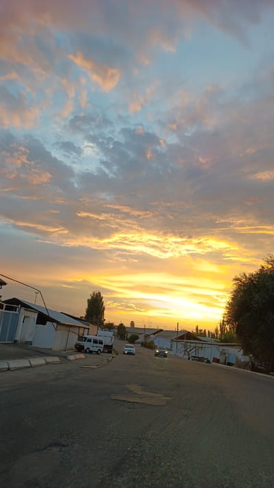 The image depicts a street scene in Yallama, Uzbekistan, during sunset. The sky is filled with dramatic clouds, transitioning from pale blue at the top to vibrant orange and yellow near the horizon, illuminated by the setting sun. The street is paved with asphalt and shows signs of wear, with some potholes visible.
On the left side of the street, there are several single-story buildings with white and blue facades. A white van is parked near these buildings. Further down the street, two cars are moving away from the viewer. The building on the right side of the street appears to be a row of structures with corrugated metal roofs and open spaces. A large, dark green tree is prominent on the right side of the image, with more trees visible in the distance. The overall impression is of a residential or semi-industrial area bathed in the warm light of dusk.