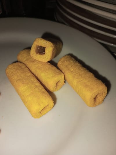 Scene in Melakudikadu, India. Five yellow-orange, cylindrical snack items are arranged on a white, slightly curved plate. One snack, situated at the upper center, is broken or cut transversely, exposing a square-shaped hollow interior filled with a dark reddish-brown substance. The other four snacks are intact, exhibiting circular or slightly oval hollow openings at their ends. The snacks possess a textured, possibly seasoned or dusted, surface. In the upper right background, a stack of two or more plates with distinct brown horizontal stripes is partially visible against an out-of-focus, dark environment. The scene is illuminated by direct light, casting shadows from the snacks onto the white plate.