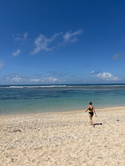 A photograph taken in Kutuh, Indonesia, shows a beach scene under a blue sky with scattered clouds. The ocean runs horizontally across the middle of the frame, transitioning from a light blue near the shore to a darker blue further out. On the right side of the image, a sandy beach covers the bottom half, appearing light tan and dotted with small shells. A person wearing a black sports bra and shorts walks on the sand, leaving footprints. Their shadow is visible on the beach next to them. The horizon line is mostly level, with the sky and ocean filling the left side of the image.