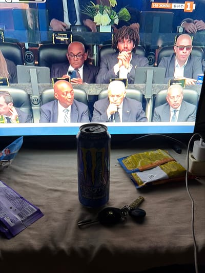The image shows a television screen displaying a broadcast, likely of a formal event or a sporting match, with several individuals seated in what appears to be a stadium or conference setting. The people on screen are dressed in suits, suggesting a professional or official capacity. In the foreground, resting on a textured surface, are several objects: a can of Monster Energy drink, two packets of snacks, a set of keys, and a portion of a purple wrapper from a confectionery item. The lighting suggests an indoor environment, possibly a living room or den, where the television is being watched. There is no specific indication of the location being in the city of Gorni Dabnik, Bulgaria, or any other specific geographic location.