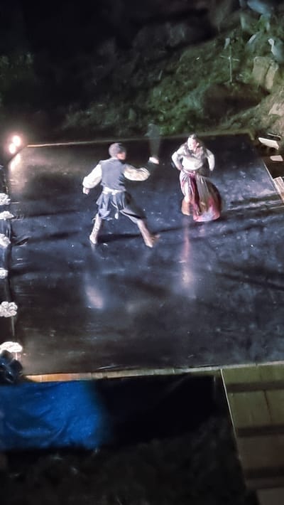 The image shows two people lying on a black surface. The person on the left appears to be a woman wearing a dress and a head covering. The person on the right appears to be a man in a tunic and breeches. Both are lying on their backs, with their legs extended. There is a faint, glowing pinkish light emanating from between them, suggesting some kind of magical or otherworldly effect.

The setting appears to be outdoors, possibly at night, given the darkness and the presence of artificial lights at the bottom of the image. The ground around the black surface is rocky and covered in greenery, suggesting a natural, possibly rugged environment. There is also a wooden structure visible in the background.

The overall impression is one of stillness and mystery. The poses of the individuals suggest they might be unconscious or incapacitated, and the glowing light adds an element of intrigue. The time of day is likely night or dusk due to the low light conditions and the visible artificial lights. The location context of Sant Joan Les Fonts, Spain, known for its volcanic landscapes and natural beauty, is consistent with the rocky and green surroundings depicted. There is no visible text in the image.