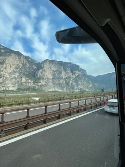 The image shows a view from inside a vehicle, likely a car or bus, traveling on a roadway in the vicinity of Mezzocorona, Italy. A mountain dominates the view, displaying a steep, rocky face with visible geological features and some vegetation. The sky is visible with scattered clouds. Adjacent to the roadway is a structure with regularly spaced vertical supports, potentially a noise barrier or a fence. Another vehicle, with the license plate "618 5889", is partially visible in the upper right corner of the frame. The interior of the vehicle frames the scene, showing the rearview mirror and portions of the car's interior.