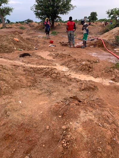 The image depicts an outdoor scene in the city of Buga da Gindi, Nigeria, characterized by a mining or excavation site. The terrain is primarily composed of reddish-brown soil and earth, with several mounds and excavated pits visible. In the foreground, the ground is uneven and appears damp in places, with some muddy areas.

Several individuals are present in the scene, engaged in various activities. In the mid-ground, three men are prominently featured. One man, wearing a red t-shirt and blue jeans, stands with his hands on his hips. Another man, in a green and white striped t-shirt and shorts, is adjusting something around his waist. A third man, wearing a blue t-shirt and dark shorts, stands with his hands on his hips and has a yellow cloth on his head. Further in the background, more individuals are scattered across the site, some appearing to be working.

Various objects are present, including what appears to be a red bucket near one of the men, discarded plastic bottles and bags scattered on the ground, and some lengths of tubing, one of which is bright orange and appears to be carrying water. The environment is open, with scattered trees and vegetation visible in the distance, and a cloudy sky overhead. The overall impression is one of active, potentially labor-intensive work in a natural, undeveloped setting.