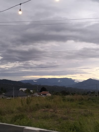The media file captures a serene landscape scene in Panteraya, Indonesia, likely at dusk or dawn due to the soft, diffused light and cloudy sky. The foreground features a rough, greyish concrete barrier or curb. Beyond this, a field of tall, dry grass stretches across the middle ground, interspersed with some greener patches. Several residential buildings with different roof colors and styles are visible within this grassy area, suggesting a semi-rural or developing community.

In the background, a series of rolling hills and mountains rise, their forms softened by the atmospheric conditions. Clouds cling to the slopes of some of the mountains, adding depth and a sense of mystery to the vista. The sky above is predominantly overcast with heavy grey clouds, but hints of lighter hues, possibly from the setting or rising sun, can be seen near the horizon between the mountain ranges.

Draped across the top of the image are two thin wires, with a single light bulb hanging from the left wire. The bulb is lit, casting a warm glow against the cooler tones of the sky and clouds. Another faint light source is visible higher up and to the right, possibly another unlit bulb or a distant light. The overall mood evoked by the image is one of tranquility and natural beauty, with a touch of rustic charm from the village nestled within the landscape. There is no visible text in the image.