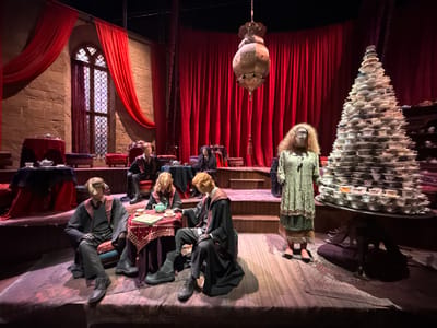 This media file depicts a theatrical or museum exhibit designed to evoke a magical or historical setting. The scene features several mannequins dressed in what appear to be school uniforms, suggesting a connection to a fictional academy. One mannequin, positioned centrally, wears a dark robe with red trim and sits at a table laden with a teapot and cups, indicating a tea ceremony or gathering. To its left, another mannequin in similar attire is also seated at a table. A third mannequin to the right, standing, is adorned in an ornate green dress with fringed layers and wears glasses.
Behind the figures, the setting is characterized by rich red drapery covering the walls and a tall, arched window with intricate panes. Stone textures are visible, contributing to an old-world ambiance. A prominent object in the background is a large, metallic lantern hanging from the ceiling. To the far right, a towering, conical arrangement of stacked teacups and saucers draws attention, creating a whimsical and slightly precarious display. The floor is covered with patterned rugs.
The lighting suggests an indoor setting, possibly during a simulated evening or a dimly lit day, enhancing the theatrical mood. The overall atmosphere feels formal and perhaps slightly mysterious, consistent with a scene from a fantasy narrative. There is no visible text within the image. The location context of Watford, United Kingdom, might suggest this is part of a known attraction or studio tour, possibly related to a film production.
