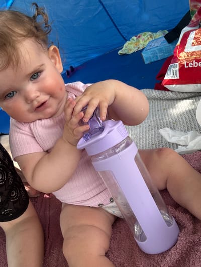 A close-up, overhead shot shows a baby with blue eyes and curly blonde hair being held. The baby is wearing a light pink ribbed onesie and has a bottle with a purple cap in its hands. The bottle is transparent and appears to contain liquid. The baby's face is turned towards the camera, and it has a slight smile. Some red marks are visible on the baby's cheeks.

The baby is lying on a textured pink blanket. To the left of the baby, there is a bright blue surface, likely a mat or tarp, and a red and white bag with some text. Part of the text on the bag is visible: "Lebe" and a barcode with numbers. There is also a small blue box and a piece of fabric with a floral pattern on the blue surface.

The overall scene suggests an outdoor setting, possibly a picnic or a day at the park, given the presence of a mat or tarp and the casual attire. The lighting appears to be natural daylight. The baby seems content and engaged with the bottle. The context provided indicates the location is Giessen, Germany.