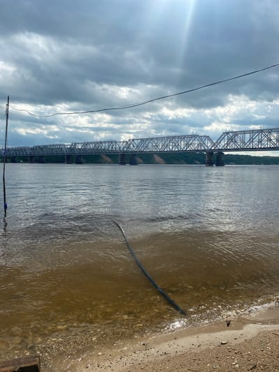 The image depicts a body of water, likely a river, under a cloudy sky. A large, silver metal truss bridge spans across the water, stretching from the left side of the frame towards the right background. The bridge appears to be carrying a railway line. On the right side of the image, there is a sandy bank with some debris and a dark, curved object, possibly a pipe or hose, partially submerged in the water near the shore. In the sky, there are dramatic, white clouds against a blue-grey background, suggesting overcast or rapidly changing weather. Sunlight pierces through the clouds on the far left, creating a lens flare effect. A few power lines are visible in the foreground, stretching across the frame from bottom left to upper right. The water is choppy with visible ripples and reflections, indicating some movement or wind. The scene appears to be set during the daytime, given the lighting conditions and sky. The location is identified as Zelenodol'sk, Russia, which implies the bridge is a significant piece of infrastructure in that area, likely a crossing over a substantial river. There is no visible text in the image. The overall mood is somewhat dramatic due to the sky and the imposing structure of the bridge.
