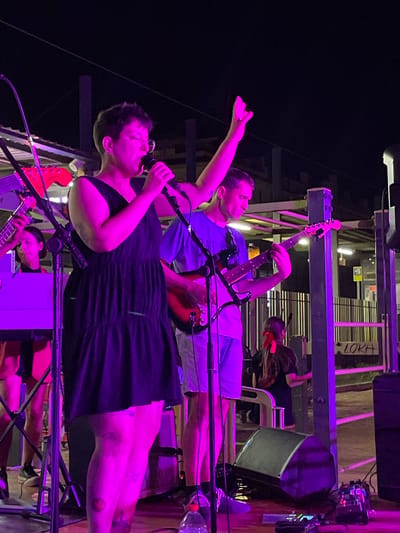 In Canet de Mar, Spain, at night, a band performs on an outdoor stage illuminated by purple lights. The lead singer, a woman with short dark hair and glasses, wears a black dress and sings into a microphone, her left arm raised. Beside her, a man in a blue t-shirt plays an electric guitar, his gaze directed downwards. In the background, another person plays a keyboard, and further back, a person with a red bow in their hair stands near a fence with "LOKA" spray-painted on it. Stage equipment, including microphones, guitars, speakers, and effects pedals, are visible on the stage. The setting appears to be an open area with some urban structures visible behind the stage.