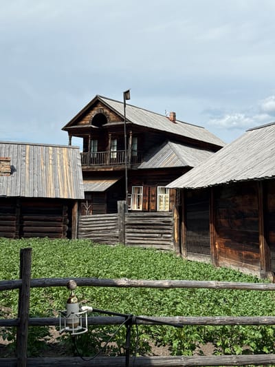 This image depicts an outdoor scene in the village of Shushenskoye, Russia, likely at an ethnographic museum or historical recreation. In the foreground, a rustic wooden fence made of rough-hewn logs stretches across the frame. Attached to the fence, a vintage-style microphone with a gold grille and white body is mounted on a stand, with cables extending out of frame. Behind the fence, a field of green plants, possibly potato plants, is visible. The background features several traditional wooden buildings constructed from logs. The central building is a two-story structure with a pitched roof, a prominent balcony with a railing on the upper level, and arched detailing above the balcony. Adjacent to it are lower, single-story wooden structures. The architecture suggests a historical setting, characteristic of rural Russian settlements. The sky above is overcast with scattered clouds. The overall impression is one of a preserved historical rural landscape.