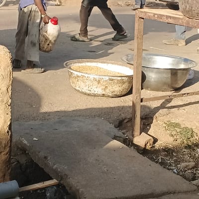 A man holding a red-capped container stands beside a large, weathered bowl filled with grain. Another bowl, shiny silver, sits nearby. These are on the pavement next to a metal frame. Pedestrians walk by. The scene is a bustling, likely outdoor market or food preparation area. A concrete slab covers a drain or ditch. The area appears dusty and well-used.