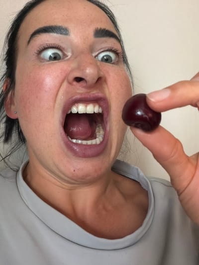 A woman is holding a cherry with her fingers, bringing it towards her open mouth. Her eyes are wide, and her facial expression suggests surprise or anticipation. The background is a plain, light-colored wall. The scene is indoors, with no specific identifiable features of Koltushi, Russia.