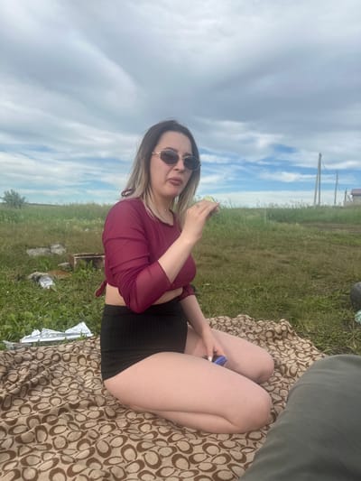 A young woman with blonde-streaked hair and sunglasses sits on a patterned blanket outdoors, holding a piece of food to her mouth. She wears a maroon sheer mesh top and black shorts. The background is a grassy field under a cloudy sky, with scattered debris, including a wooden box and foil wrappers, on the left. Power lines and a distant building are visible on the right. The setting is consistent with an outdoor recreational area in Ozyorny, Russia.