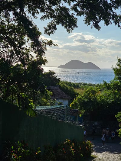 The image captures a coastal scene in Armação dos Búzios, Brazil, framed by lush foliage. A shimmering body of water stretches towards a mountainous silhouette on the horizon, partially obscured by clouds. Power lines run horizontally across the water. A structure, possibly a house, is visible amidst the greenery. On the right, people are walking on a paved pathway. The scene is bathed in sunlight.