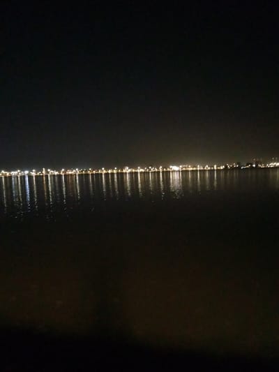 This media file captures a nighttime scene of a vast body of water, likely a bay or sea, with a distant city or town skyline illuminated by artificial lights. The upper portion of the image is dominated by a dark, starless night sky, indicating deep night. Along the horizon, a continuous line of numerous bright, warm-toned lights stretches across the frame, emanating from buildings or infrastructure on the distant shoreline.
These urban lights cast distinct, elongated reflections that shimmer and extend vertically downwards across the dark, calm surface of the water. The reflections are almost as prominent as the lights themselves, suggesting still water conditions. The foreground consists of the dark water, gradually fading into deeper shadow at the very bottom of the frame, offering no specific details.
There are no people or discernible activities taking place in the image. The setting conveys a peaceful and expansive nocturnal landscape, with the artificial glow of human settlement contrasting against the natural darkness. Given the provided context, this scene likely depicts a waterfront area in Al Khobar, Saudi Arabia, such as its Corniche, at night. No visible text is present in the image.