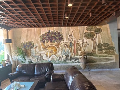 This image depicts the interior of a common area or lobby located in Melnik, Bulgaria. The dominant feature is a large, multi-panel mural spanning the rear wall, rendered in an ancient pastoral style on a textured, light-colored background.

The mural illustrates various scenes: On the left, a hooded male figure is seated, interacting with a smaller figure, while a large basket overflowing with purple and red grapes, accompanied by a black bird, is depicted behind them. Towards the center-left, a male figure reclines while playing a pipe-like instrument, with a large circular object nearby. In the center, a seated male figure with dark curly hair is accompanied by a dog. To his right, a standing male figure holding a long staff and wearing a red cloak is shown. Further right, stylized trees with layered green foliage and two horned animals resembling bulls are present. Small green bushes line the bottom of the mural on the right.

In the foreground, two dark brown leather sofas are visible. One, a two-seater, is positioned on the left, partially facing the viewer. Another sofa or a section of a larger sofa is seen on the right, also in dark brown leather. A rectangular coffee table with a dark wooden frame and a glass top sits between the sofas. On the coffee table, a small glass vase holds white hydrangea flowers, next to a wooden box labeled "DOMINO."

The ceiling is a dark brown wooden grid, forming a coffered pattern of square recesses. Multiple light fixtures are integrated into the ceiling: several cylindrical downlights (some dark brown, some metallic) and a modern pendant light with a dark drum shade near the left window.

To the left, a large window is draped with translucent, light beige curtains, revealing green foliage and trees outside. Several potted plants are placed near the window, including a tall, slender plant and smaller plants on the windowsill. The floor is covered with light-colored, square tiles, likely marble or polished stone, featuring subtle veining. On the far right, a large, terracotta-colored amphora-style urn, containing dried plant material, rests on a black metal tripod stand. A section of a blue-grey wall or pillar is visible on the far right edge of the frame.