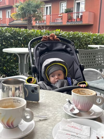 The image presents an outdoor cafe setting in Longone al Segrino, Italy. In the foreground, a light-colored outdoor table displays multiple items. On the left, a white ceramic coffee cup with a colorful gradient design is partially empty, showing "illy espresso" and "Italy Chicago" branding, resting on a white saucer with a silver spoon nearby. Adjacent to it, a silver metallic pitcher stands next to a light blue smartphone, positioned upright. To the right, a pink and white striped ceramic coffee cup, also bearing "illy espresso" text, is mostly empty on a white saucer with a spoon. Two white paper packets are visible on a small plate.

In the midground, an infant is seated in a dark grey stroller. The baby wears a grey knitted beanie with a yellow trim and a dark grey hooded jacket, looking towards the right. A small, brown, plush toy is visible near the top of the stroller. A white slatted outdoor chair is partially visible to the right of the stroller.

The background features a dense green hedge, behind which stands a reddish-brown residential building. This building has multiple white-framed windows, doors, and balconies with red railings adorned with potted plants. A large palm tree is visible in the center-left background. The sky appears overcast.