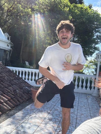 A male subject stands on an outdoor tiled terrace in San Pablo La Laguna, Guatemala, under bright daylight. He is positioned centrally, balancing on his right leg with his left leg raised. The subject has dark, curly hair and a fair complexion, displaying an open-mouthed expression with his tongue extended. He wears a light-colored t-shirt featuring a green avocado graphic on the chest and black shorts. Brown sandals are visible on his right foot. His right hand is positioned near the avocado graphic, while his left hand rests by his hip.

To the subject's left, a terracotta-tiled roof is visible. Behind him, a white balustrade separates the terrace from a dense background of green foliage and trees. A small, white, domed architectural feature with a dark railing is partially visible within the foliage on the far left. The terrace floor is composed of light-colored, patterned square tiles, and a brown terracotta plant pot is situated near the subject's left leg. Bright sunlight emanating from the upper left quadrant of the frame creates a distinct lens flare. The upper right portion of the background shows a clear blue sky with some cloud formations.