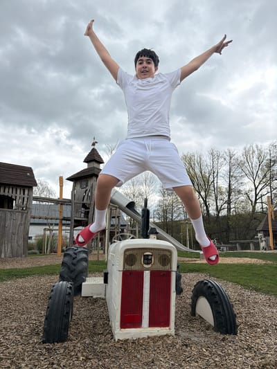 A young person wearing a white t-shirt, white shorts, white socks, and red sandals is lying across the hood of a small, white and red tractor. The person is posed with their arms spread wide, parallel to the ground, and their head is tilted towards the viewer. The tractor is situated on a surface covered in wood chips. In the background, there's a playground structure made of wood, with towers, ropes, and a slide. The sky is overcast. It's impossible to pinpoint the location in the city of Olingen, Luxembourg, from this image alone.