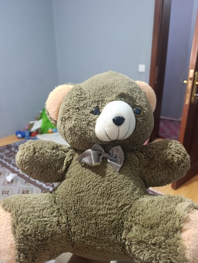 A close-up shot features a fluffy brown teddy bear, positioned centrally. The bear has a white muzzle, black button nose, and large dark eyes with a hint of reflection. It sports a gray satin bow tie at its neck. The bear's ears are lined with a lighter, peachy-pink material. The foreground is dominated by the teddy bear, with its fuzzy texture clearly visible.
The background is softly blurred, suggesting an indoor setting. To the left, a wall is visible, painted in a light gray-blue hue. A dark wooden door with brass accents stands on the right. Beneath the bear, a patterned rug is visible, and beyond that, a wooden floor. In the far background, several colorful objects, possibly toys, are scattered on a surface, with a hint of green fabric. The lighting suggests it is daytime, with natural light filtering into the room, creating a warm and casual atmosphere. The overall impression is one of a cozy, domestic environment. No text is visible in the image.