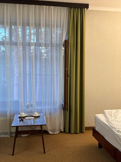 This image captures an indoor scene, likely a hotel room in Журавлево (Кемеровский), Russia. A large window is covered by two layers of curtains: sheer white curtains in the foreground and heavy olive-green curtains to the right. The white curtains reveal a blurry view of trees outside. On a small, dark wooden table in front of the window, there are two cups and saucers, four bottles of water, and two glasses. To the right of the window, a portion of a bed with white linens is visible. The walls are textured in a light beige color. The flooring is a coarse, speckled brown carpet. The scene is lit with artificial lighting, casting a soft glow on the textured wall. There are no people visible in the image.