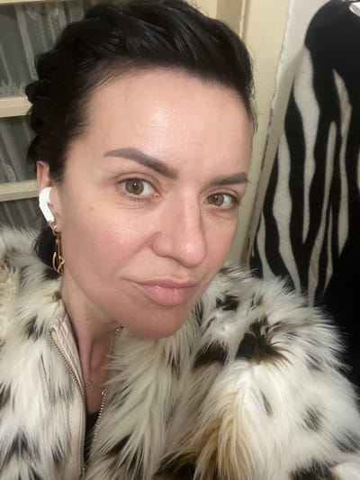 A medium close-up, chest-up portrait depicts an adult female indoors in Smederevska Palanka Municipality, Serbia. The subject, estimated to be middle-aged, has dark, slicked-back hair, light brown eyes, and is looking directly into the camera with a slight smile. She is wearing a cream-colored faux fur jacket featuring irregular dark brown or black spots, with a light-colored zipper visible on the front. A white wireless earbud is present in her left ear, beneath which a gold-toned, abstractly shaped earring hangs. A thin silver-colored necklace with a small pendant is partially visible below the jacket's opening. The background on the left side of the frame shows a light-colored surface with horizontal elements, possibly a window blind or part of a furniture piece. To the right of the subject, a black and white zebra-striped fabric or object is visible.