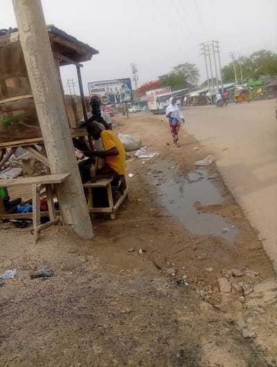 The image depicts a street scene in Aviation Quarters, Nigeria. In the foreground, a man in a yellow shirt sits on a small wooden stool, appearing to be working on something. Another man sits nearby. They are positioned near a weathered wooden structure resembling a makeshift shop or workstation. A utility pole stands adjacent to the structure.

A puddle of water with debris and trash is visible on the ground near the road. A woman in a white hijab and colorful patterned clothing walks along the side of the road. In the background, there is a road with vehicular traffic, including a Hansel truck and other vehicles. A billboard for Crestfield Metropolitan Schools is visible. Overhead power lines run across the scene. The sky is overcast.