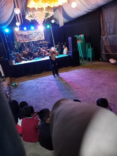The image captures an evening cultural or musical event held under a temporary tent structure in Pangalengan, Indonesia.
In the background, a raised stage, draped in black, hosts a group of approximately 10-15 male musicians. They are actively performing, playing traditional instruments such as drums (kendang) and other percussion or stringed instruments, typical of Indonesian musical ensembles. A banner, partially obscured, hangs above the stage with visible text that includes "LINGKUNG SENI," "PUSTAKA," "NYARENGAN," and "KAMPUNG," likely detailing the performing group or event. The stage is brightly illuminated by blue and green stage lights, and large speakers flank either side.
In the midground, a woman wearing a light brown hijab, black top, and black pants stands on the dirt ground, facing the stage. Her figure casts a long, colored shadow due to the stage lighting. To the right of the stage, a stack of green plastic chairs is visible against a black tarp wall that forms part of the tent.
The tent's ceiling is decorated with white and light blue fabric drapes, from which several white spherical lanterns and other decorative elements hang. The ground in front of the stage, where the woman stands, is a dirt surface, vividly colored with purple and pink hues from the stage lights.
In the immediate foreground, several children are seen from behind, sitting on the ground and seemingly watching the performance. Part of what appears to be the viewer's arm or shoulder obstructs the lower right portion of the frame. The overall atmosphere suggests an engaged community gathering or cultural celebration.