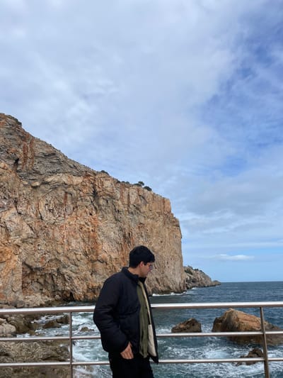The scene depicts a male individual on a coastal promenade in Torroella de Montgrí, Spain. The individual, positioned in the lower center of the frame, has dark hair and glasses, and wears a black puffer jacket over a green inner layer, with dark trousers. His right hand is in his pocket, and he is looking downward towards the water. A silver-colored metal railing, comprising two horizontal bars, separates the promenade from the ocean. To the left and extending upwards is a massive, multi-textured rocky cliff face, predominantly light brown and orange, exhibiting visible cracks and sparse green vegetation at its summit and along ledges. The agitated ocean water, dark blue-green with white wave crests, contains several large, dark rock formations near the shore and extends to the distant horizon on the right. The sky overhead features a mix of light grey cloud cover and patches of visible blue sky.