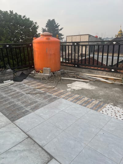 This image depicts an outdoor scene on a rooftop in Pasir Gadung, Indonesia, likely a residential area given the surrounding buildings and vegetation. In the foreground, a partially tiled patio area transitions to a concrete surface. Scattered on the concrete are various construction materials, including pipes, hoses, and coiled rope. An orange water tank is prominently positioned on the left side of the frame, resting on concrete blocks. A black metal railing runs along the edge of the rooftop, separating it from the lower buildings and trees visible in the background. The sky is overcast, suggesting it might be early morning or a cloudy day. The overall impression is of an unfinished or under-construction outdoor space.