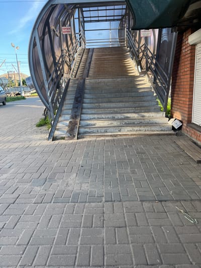 An outdoor shot shows a pedestrian overpass with stairs and a ramp leading upwards, likely in the city of Kalynivka, Ukraine. The overpass has a metal frame with a translucent curved roof, and a green mesh shade is visible at the top right. Red brickwork forms part of the structure on the right, adjacent to a white roll-up door. The ground in front of the overpass is paved with grey interlocking brick pavers. In the distance, streetlights, power lines, and some industrial or agricultural structures are visible. Two signs are attached to the overpass: one red sign reads "ТОРГІВЛЯ ЗАБОРОНЕНА!" (Trading is forbidden!), and another white sign states "НЕ СМІТТЯ У СВОЄМУ ЗОВНІШНЬОМУ ВДІ" (Do not litter in your external appearance). A white plastic box with a lid is on the ground near the brick wall and stairs. A cigarette butt and a piece of green wire are on the paved ground in the foreground. The lighting suggests it is daytime, with shadows cast by the overpass on the pavement.
