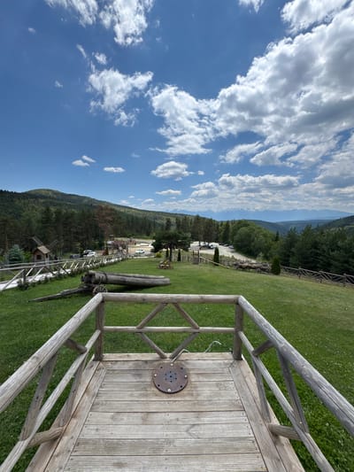 The image depicts a panoramic view of a rural landscape with a wooden observation deck in the foreground, overlooking a grassy hill. Beyond the hill, there is a cluster of buildings including a windmill, a parking lot with several cars, and in the distance, a forest-covered mountain range under a partly cloudy blue sky. The wooden deck features a railing made of logs and planks, and a circular metal plate is embedded in its surface. On the grassy slope below the deck, there is an old wooden cart with large wheels and a cannon made of logs, suggesting a historical or themed attraction. The scene is set in Yurukovo, Bulgaria, and appears to be a daytime outdoor setting with lush green vegetation and a clear sky.