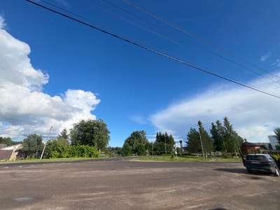 The image depicts a wide asphalt road in the city of Serebryansky, Russia. The sky is a vibrant blue with scattered white clouds, and several power lines stretch across the upper portion of the frame. On the left side of the road, a house with a brown metal roof is visible, partially obscured by trees and foliage. A utility pole leans precariously near the house.
The right side of the road features more greenery, with various trees and bushes. A dark gray Kia Sportage is parked on the shoulder, with its driver's side door open. In the background, a small brick church with a red roof and a cross on top is visible, along with other buildings and more trees. The overall scene suggests a quiet, semi-rural area within the city.