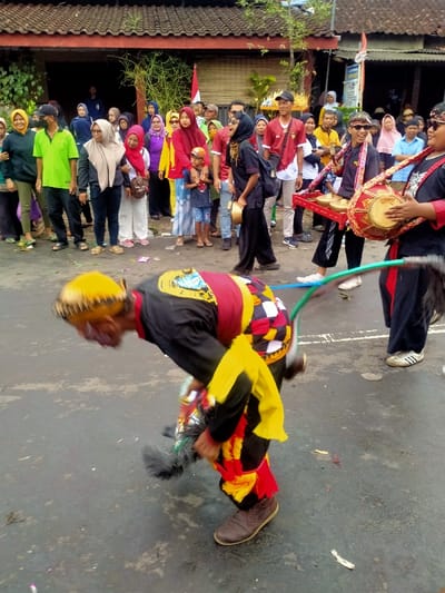 An outdoor cultural performance is depicted on a paved street in Mojogedang, Indonesia. In the foreground, a male performer, blurred due to motion, is bent forward at the waist. He wears a yellow head covering, a dark long-sleeved shirt with a red and black patterned sash, and black pants adorned with red and yellow fabric strips at the ankles, holding a dark, feathery prop. To the right, a male musician holds a red traditional kendang drum. Further left, another male musician, wearing sunglasses, carries a red tray with multiple gold-colored bonang gongs suspended from his shoulder. Both musicians wear dark clothing with red accents and white sneakers. In the background, a diverse crowd of approximately 25-30 individuals, including men, women (many wearing hijabs), and children, observe the event from the side of the street. They are dressed in casual attire. Traditional buildings with brown tiled roofs line the background, and an Indonesian flag is visible on a pole. The street surface is dark gray asphalt, showing minor debris.