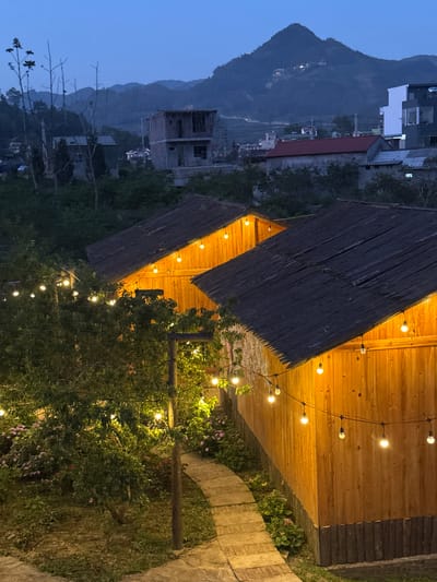 The photograph depicts an outdoor scene in Bắc Hà, Vietnam, at dusk. A cluster of thatched-roof structures, possibly part of a restaurant or homestay, is illuminated by strings of small, warm-toned lights. Wooden walls and walkways are visible. Dense foliage and greenery fill the foreground and midground, partially obscuring the structures. In the distance, a mountain range and residential buildings are silhouetted against the evening sky. The image captures the atmosphere of a rural or semi-rural location with a mix of natural and built elements.