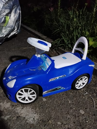A blue and white toy car is situated on dark, uneven ground, possibly asphalt or packed dirt, with some gravel and debris scattered around. Tall, dark green foliage forms a dense background, suggesting an outdoor setting, possibly a yard or park. A silver, crinkled object, potentially a tarp or protective cover, is visible on the left side of the frame. The toy car, designed to resemble a sports car, features white accents on the front, a white seat, and white wheels with a spoke design. A white handle extends from the rear of the car. The car appears slightly weathered, with some scuff marks and faded stickers on its body. The lighting suggests it is nighttime or dusk, with artificial light illuminating the scene. The location is identified as Shchastia, Ukraine.