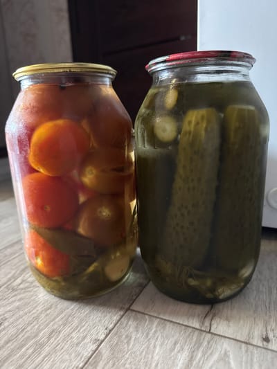 The image shows two glass jars on a wooden floor. The jar on the left contains red cherry tomatoes and some bay leaves. The jar on the right contains several whole pickles. Both jars are sealed with metal lids. The background is blurred, but appears to be an indoor setting. This image is not in Kandry, Russia, as there is no indication of the location.