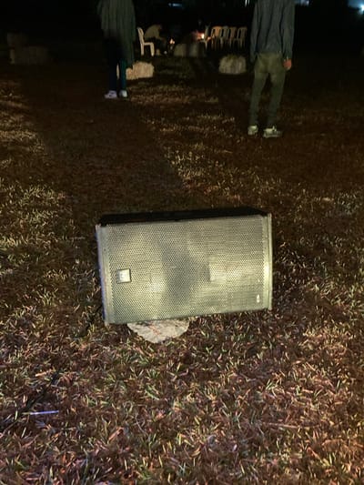 The image depicts an outdoor nighttime scene in Murera ward, Kenya. In the foreground, a black, rectangular public address (PA) speaker with a gray metal mesh grille rests horizontally on a patchy grass surface, supported by a light-colored object underneath its center. A small rectangular emblem is visible on the left side of the speaker's grille. In the mid-ground, two individuals are standing on the grass; the person on the left wears dark trousers and white athletic shoes, while the person on the right wears dark trousers and light-colored athletic shoes. In the background, several white plastic chairs are arranged on the grass, some appearing occupied. A small, glowing light source, consistent with a campfire, is visible among the chairs. Multiple irregularly shaped, light-colored objects, resembling hay bales or bundled materials, are scattered across the grass. The environment is dimly illuminated by artificial light, casting visible shadows.