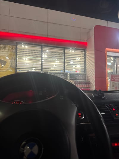 The image shows the interior of a BMW car, specifically the steering wheel with the BMW logo, the dashboard with a start/stop engine button, and the driver's view through the windshield. The location appears to be in front of a building with a red facade and large vertical windows, likely a shop or commercial establishment. The scene suggests the car is parked in front of the building.
