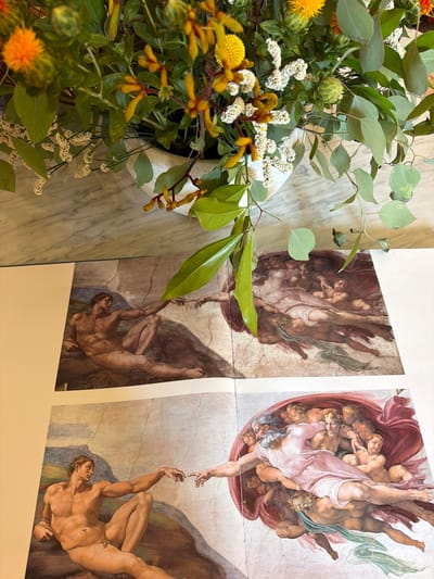 This media file showcases a close-up, angled view of two unframed prints of Michelangelo's "The Creation of Adam" from the Sistine Chapel ceiling. The prints are laid out side-by-side, with a slight overlap or gap between them. The left print depicts the figures of God reaching out to Adam, while the right print shows Adam reaching back towards God. Both prints are detailed reproductions, featuring the characteristic cracked plaster texture of the original fresco.

In the foreground, to the left of the prints, is a bouquet of greenery and yellow and white flowers. The bouquet is arranged in a pale, textured pot, and some of the leaves and branches extend over the prints, partially obscuring them. The flowers appear to be a mix of yellow thistles, small white blossoms, and eucalyptus leaves.

The background is a marble surface, suggesting an indoor setting, possibly a home or studio. The lighting appears to be natural and soft, indicating daytime. There is no visible text within the media file. The arrangement of the flowers and the presentation of the art prints suggest a decorative or artistic display. Given the location context of Fiumicino, Italy, the prints likely represent artistic appreciation or decoration in an Italian setting.