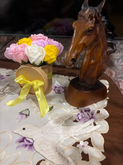 A close-up, slightly elevated shot showcases a table arrangement featuring a bouquet of colorful soap roses in a circular cardboard container tied with a yellow ribbon, a wooden sculpture of a horse's head, and an embroidered placemat with a bird and floral motifs. The arrangement is placed on a wooden table, with a dark background that appears to be a television screen and possibly wallpaper behind it. The scene is dimly lit, with the focus primarily on the objects in the foreground. The overall impression is one of decorative tabletop display.