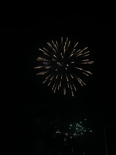 This is a photograph of a nighttime fireworks display over the city of Castelló d'Empúries, Spain. The primary subject is a large, golden-yellow burst of fireworks dominating the upper portion of the frame. The fireworks are radiating outwards with thin, bright streaks against a black sky. Below this main display, a secondary, smaller cluster of green and pink fireworks is visible. The background is entirely black, indicating a night scene, with the faint suggestion of distant, possibly urban, lights at the very bottom. The image is composed to emphasize the fireworks against the dark sky.