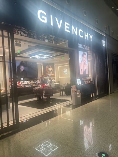 The image shows the exterior of a Givenchy store, likely located in a shopping mall in Guangzhou City, China. The store features a large, illuminated "GIVENCHY" sign on the left side of the frame. Inside the store, several display windows are visible, showcasing perfumes and makeup products. Large advertisements featuring models are displayed on screens. The store's interior design appears modern and luxurious, with clean lines and bright lighting. The floor of the mall is tiled and reflects the store's lighting and some of the advertisements. In the distance, a green directional arrow sign is visible on the floor, indicating a pathway. The overall impression is that of a high-end retail environment, likely during evening hours given the artificial lighting. There are no visible people in the foreground, but the advertisements suggest a focus on fashion and beauty. The text "GIVENCHY" is clearly visible on the main sign and as a logo inside the store.