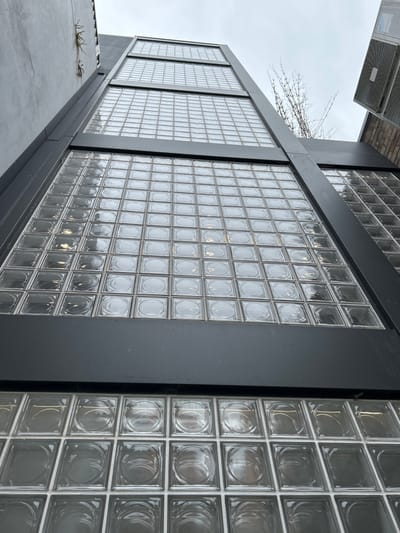A low-angle shot focuses on a building exterior featuring multiple sections of glass blocks set within dark gray frames. Each block displays a subtle circular pattern. The perspective emphasizes the height of the building and its architectural details. A faint tree branch is visible against a cloudy sky in the background. The building appears to be a modern design, utilizing glass blocks as a prominent element. An air conditioning unit is on the top left.