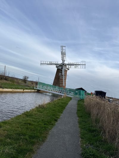 The image depicts Horsey Windpump in North Norfolk, United Kingdom, on an overcast day. A prominent brick-built windpump with four skeletal sails, devoid of fabric, stands in the midground, its base partially obscured by a temporary white metal gangway or bridge extending from the right bank across a dark body of water (likely a dyke or canal). This gangway is lined with light green netting. To the immediate right of the windpump, several temporary structures, including a dark-colored shed and other construction materials, are visible. In the foreground, a dark grey gravel path curves from the bottom right towards the center, bordered by green grass on its left and tall, dry reeds or grass on its right. The water body runs parallel to the path on the left, reflecting the muted sky. The background reveals a flat, sparsely vegetated landscape with distant trees and utility poles under a light grey, cloudy sky. No individuals are present in the scene.